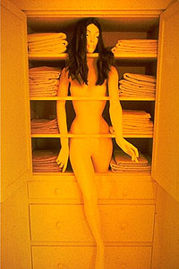 a yellow-tinted photograph of a closet with a mannequin inside. there are towels surrounding the mannequin's centered body, and she is nude, hands loosely at her sides, hands outstretched. She has long dark hair surrounding her made-up face, and a single leg emerges from her closet. The shelves separate er at her neck, under her breasts, at her torso, and above her pelvis and legs.