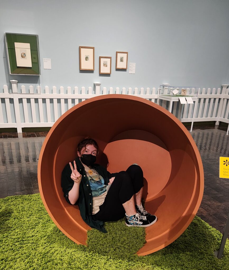 a person with hair in a ponytail and a grin sits inside a large flower pot. They hold up a peace sign, and rest their feet on the green carpet extending out. 