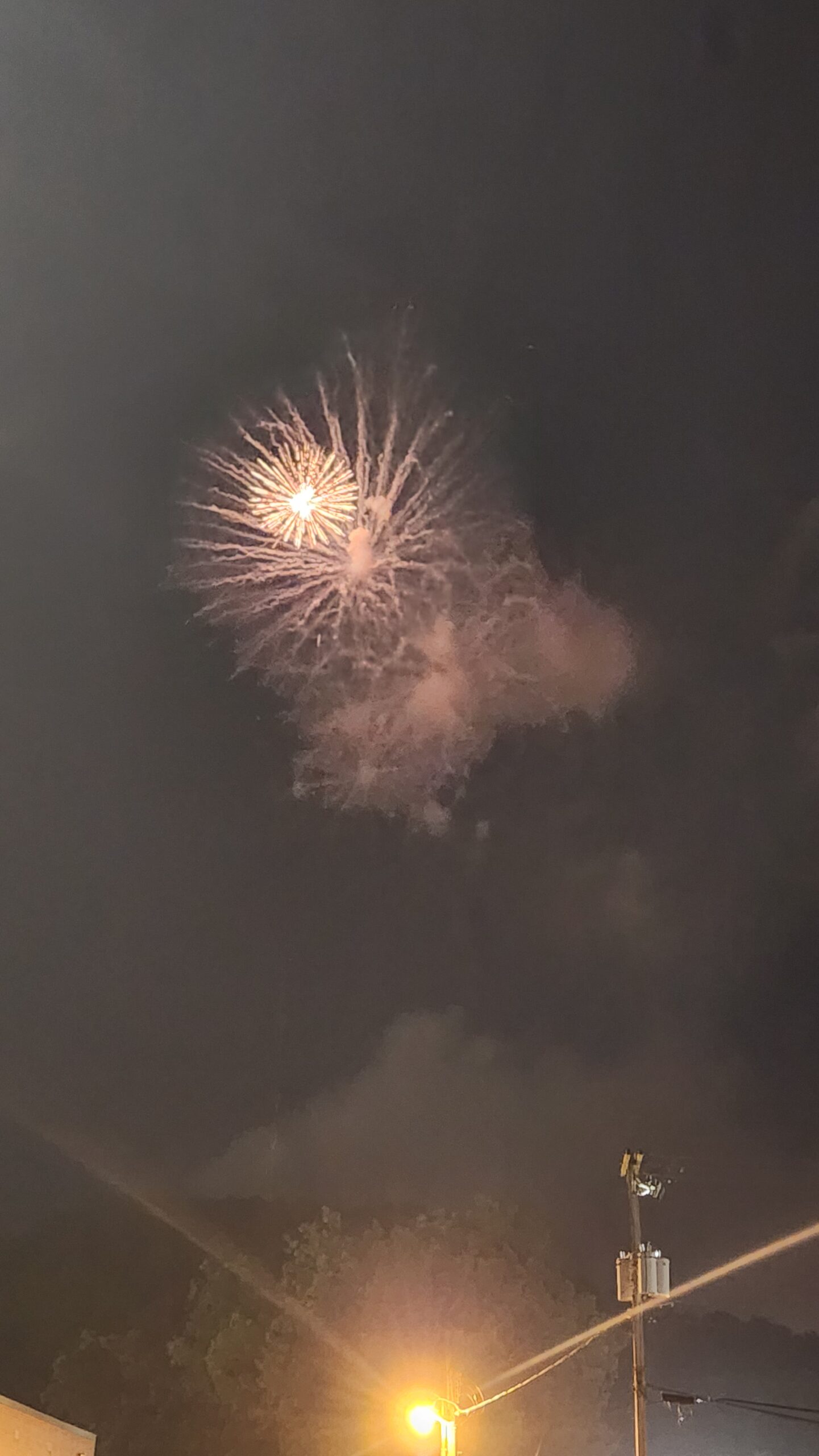 A smoky night sky, illuminated by a yellow firework exploding with pinpoints of light, highlighting smoke and clouds