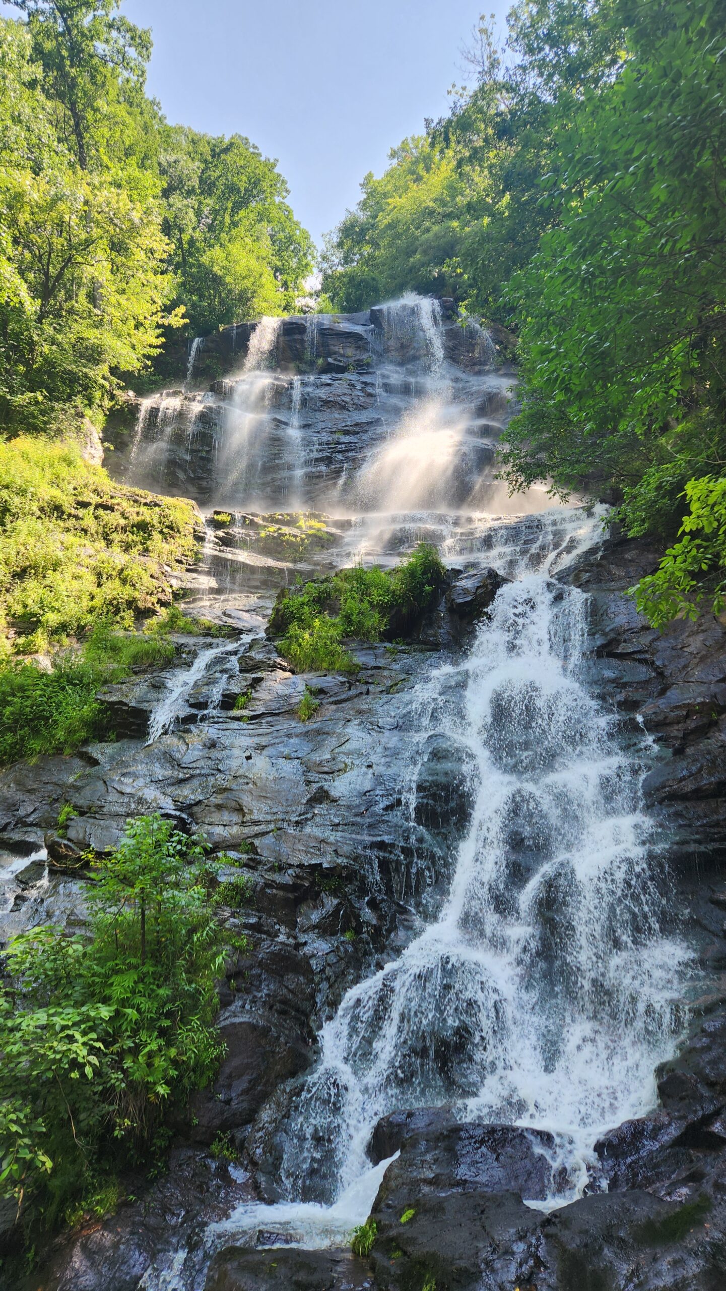 a waterfall cascading down the stones, back and forth with water and mist catching the light. Green trees and leaves are illuminated, ranging from light green to bright yellow in the sun.