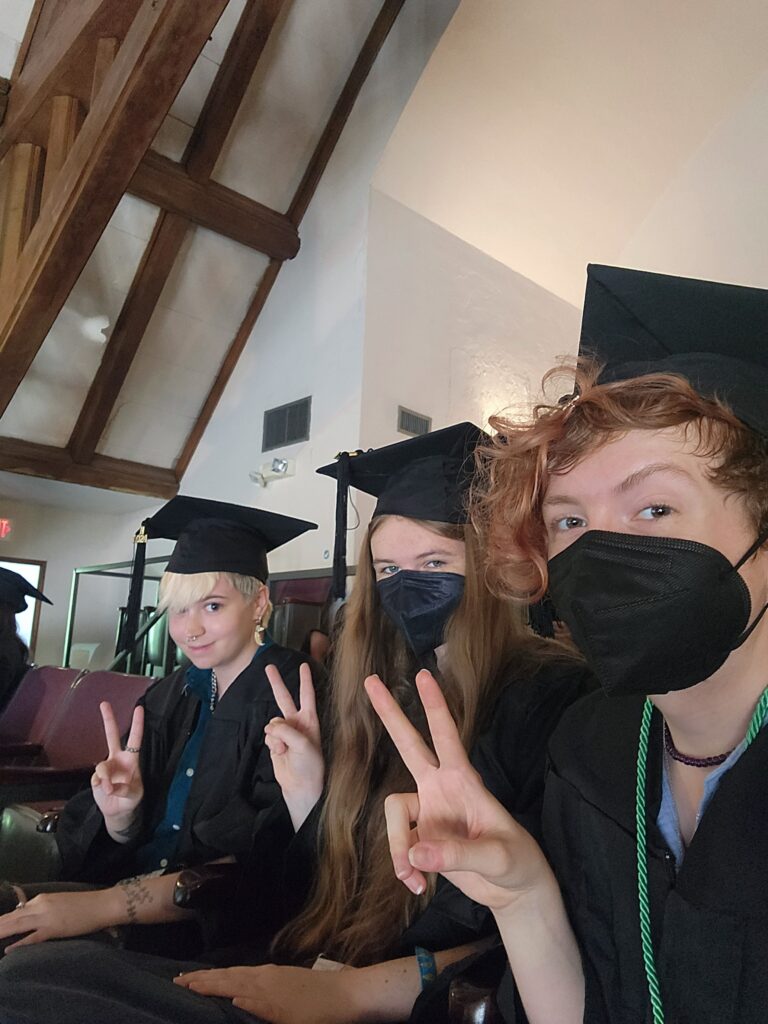 three individuals have on graduation garb, including cap and gown. From left to right, the people have short blond hair, longer brown-blonde hair and a black face mask, and short curly red-brown hair and a black face mask. 