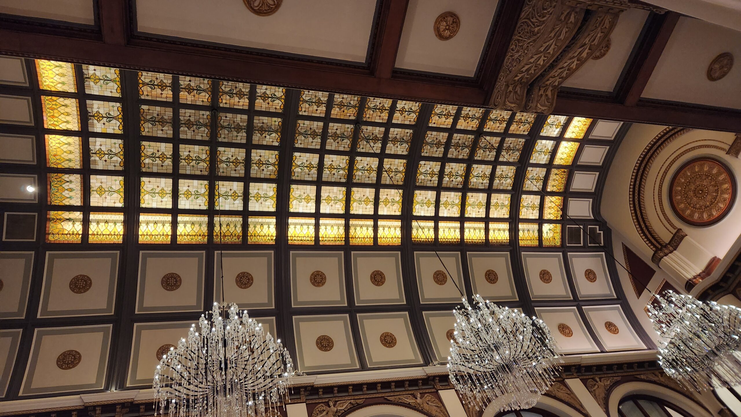 A photo looking straight up, stained glass windows in a grid on the ceiling emanating warm yellow light, chandeliers dangling below.