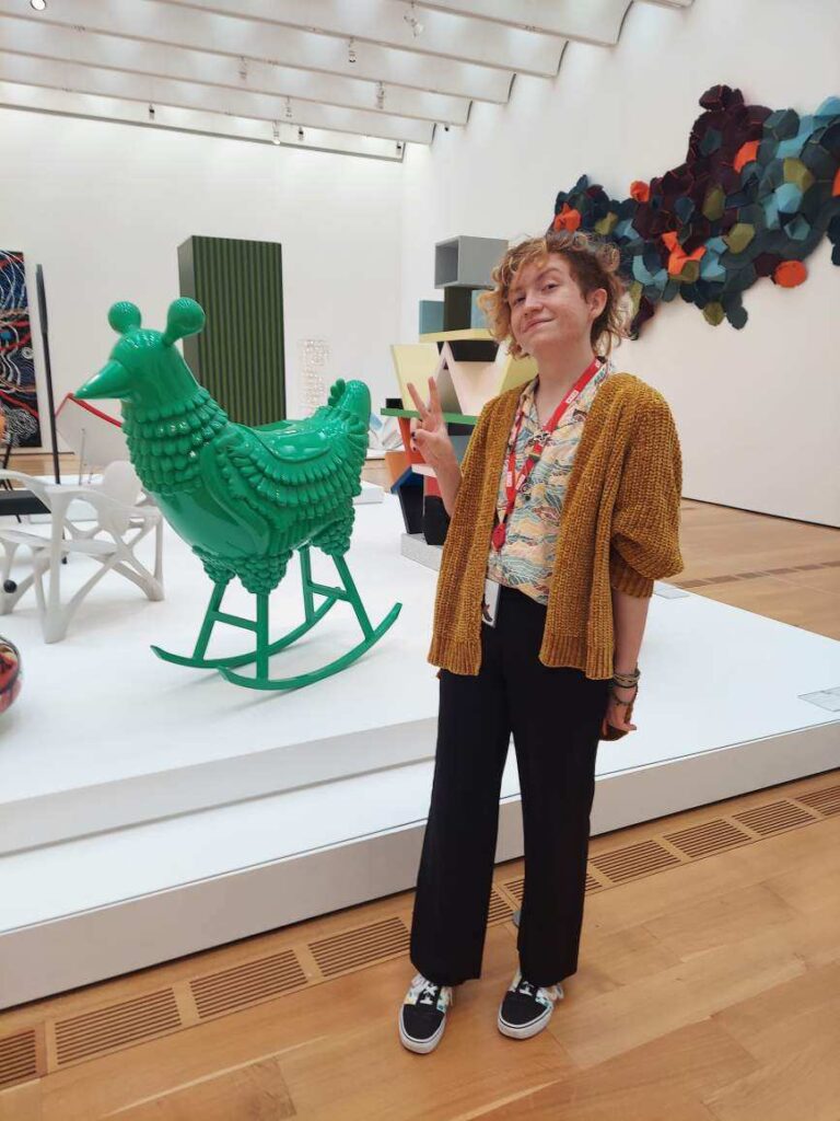 A person with short, curly hair stands in front of a chicken rocking horse sculpture which is bright green, holding up a peace sign. They wear a colorful button up, a yellow sweater, and black pants.