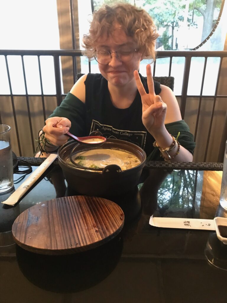 a person with short, curly reddish hair wears a black tank top with a design on the front and a green shirt, draped around their shoulders. They appear to be laughing, an are holding up a peace sign as their other hand holds up a spoonful of soup from a large pot in front of them.