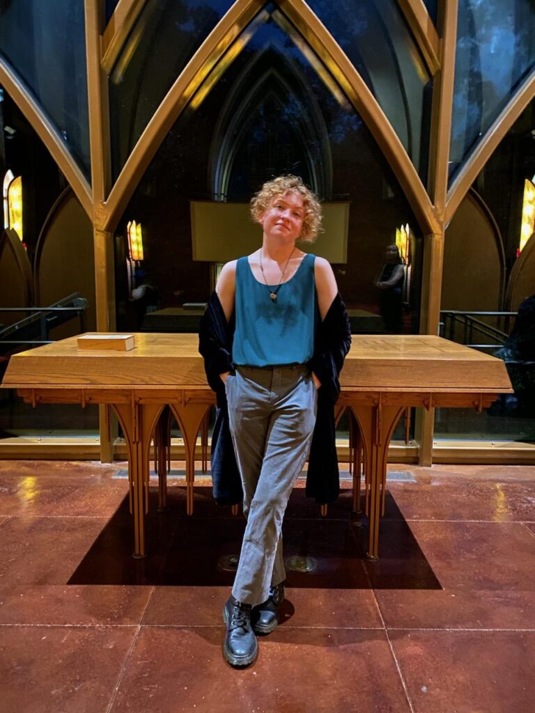 A person with short blond-red hair and a green tank top, grey pants, and boots and a black velvet wrap stands with their legs slightly crossed. They lean on a table with darkened windows in the background. 